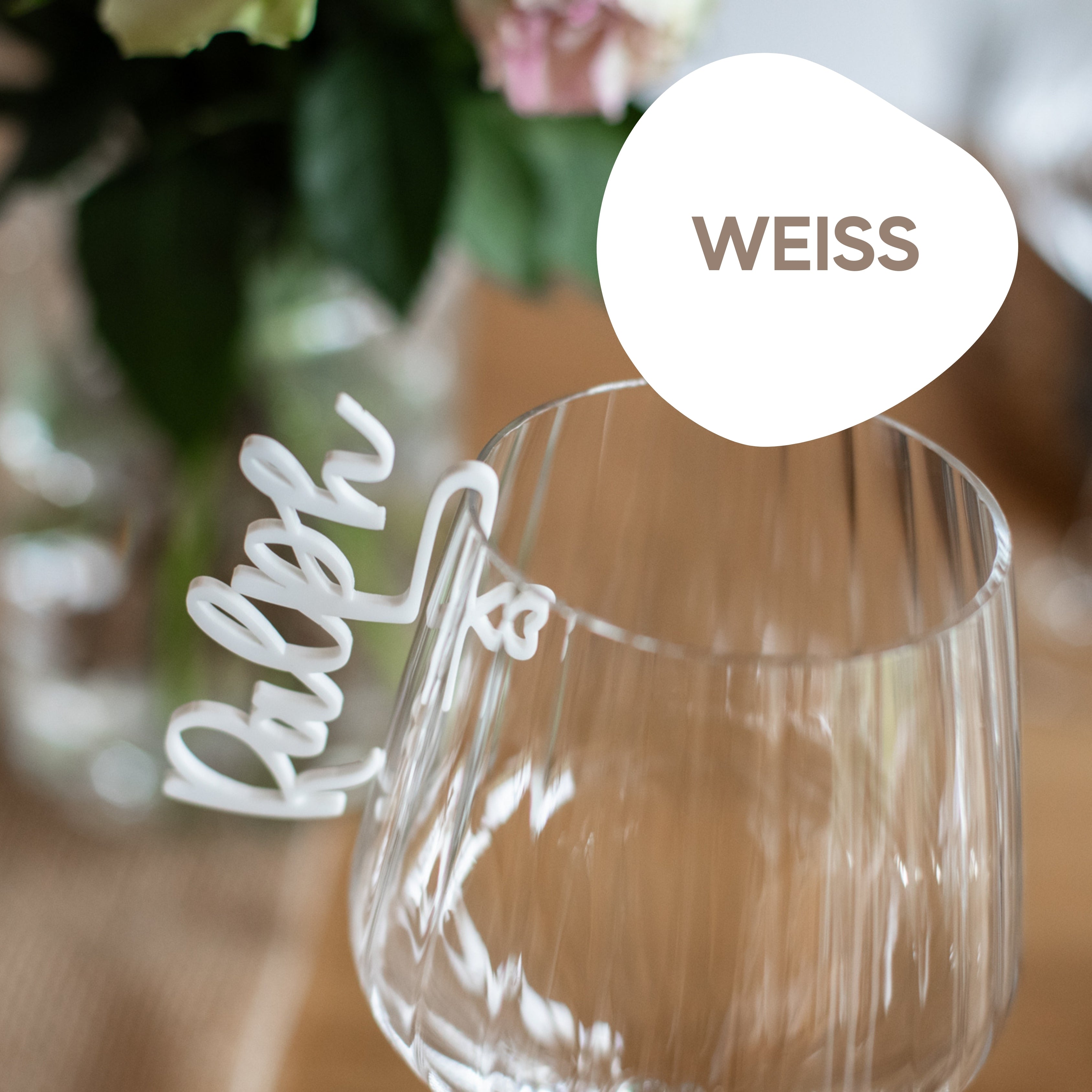 Ein Weinglas ist elegant mit einem „Weiss“-Etikett und einem „Ralph“-Clip versehen, dahinter sind verschwommene Blumen zu sehen. Ideal in Kombination mit dem personalisierten Glasmarkierer aus Acryl von laserhelden für eine unvergessliche Note.