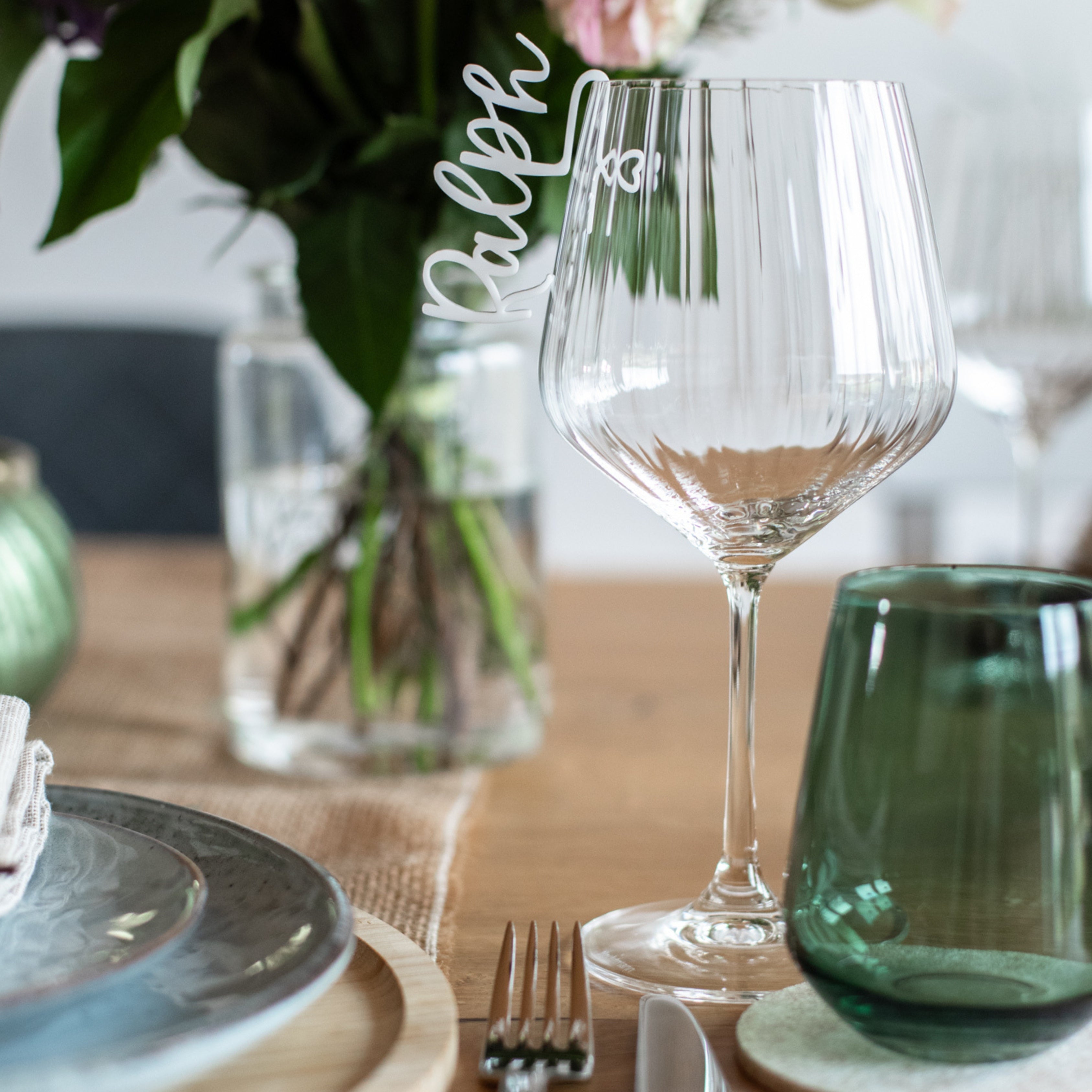 Gestalten Sie eine elegante Tischdekoration mit einem personalisierten Weinglas mit der Aufschrift „Ralph“, einem grünen Glas und einer Vase mit Blumen. Fügen Sie mit Gedecken aus dem personalisierten Glasmarkierer aus Acryl von laserhelden Charme hinzu und verstärken Sie zusammen mit dem Tischgeschirr die intime Atmosphäre.