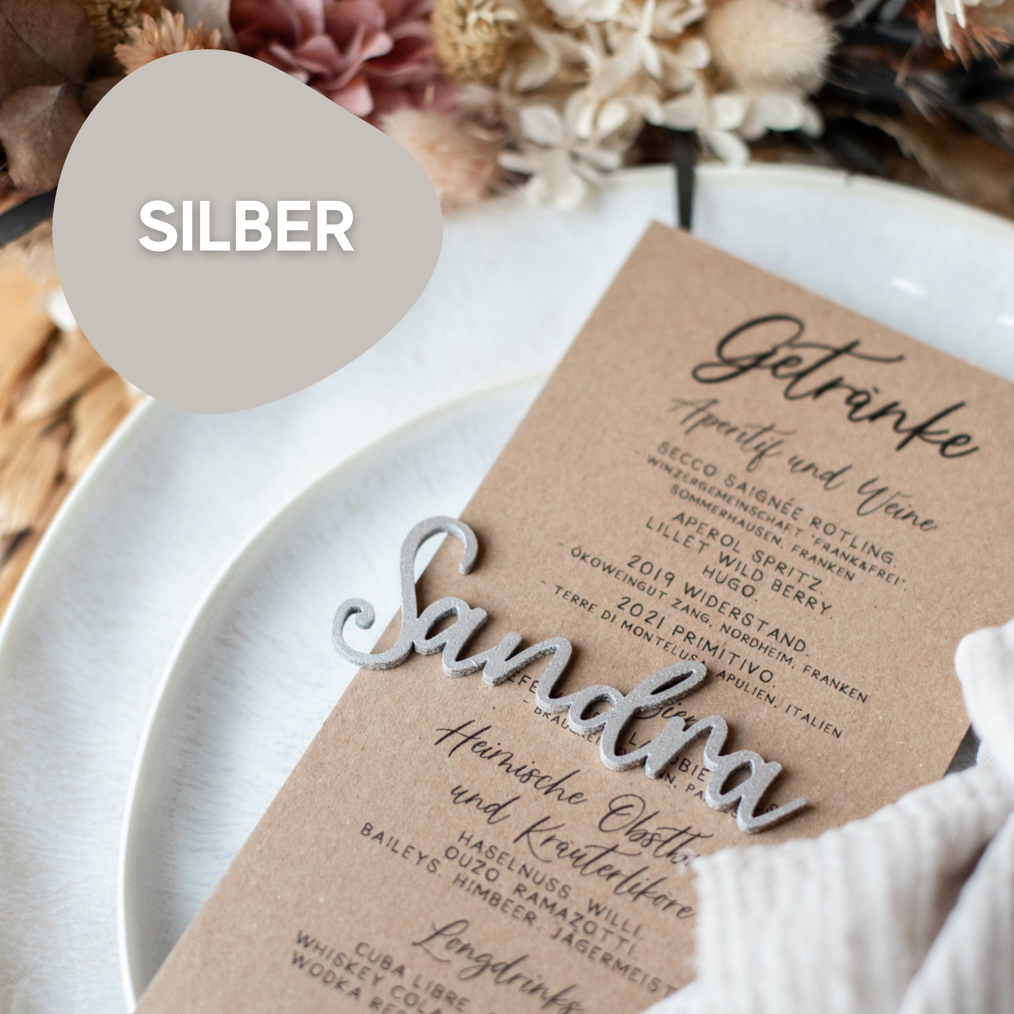 Eine Nahaufnahme eines Hochzeits-Tischkarten-Schriftzugs aus Holz in Silber von Laserhelden mit „Sandra“, umgeben von getrockneten Blumen auf einem weißen Teller, wodurch eine exquisite handgefertigte Tischdekoration entsteht.
