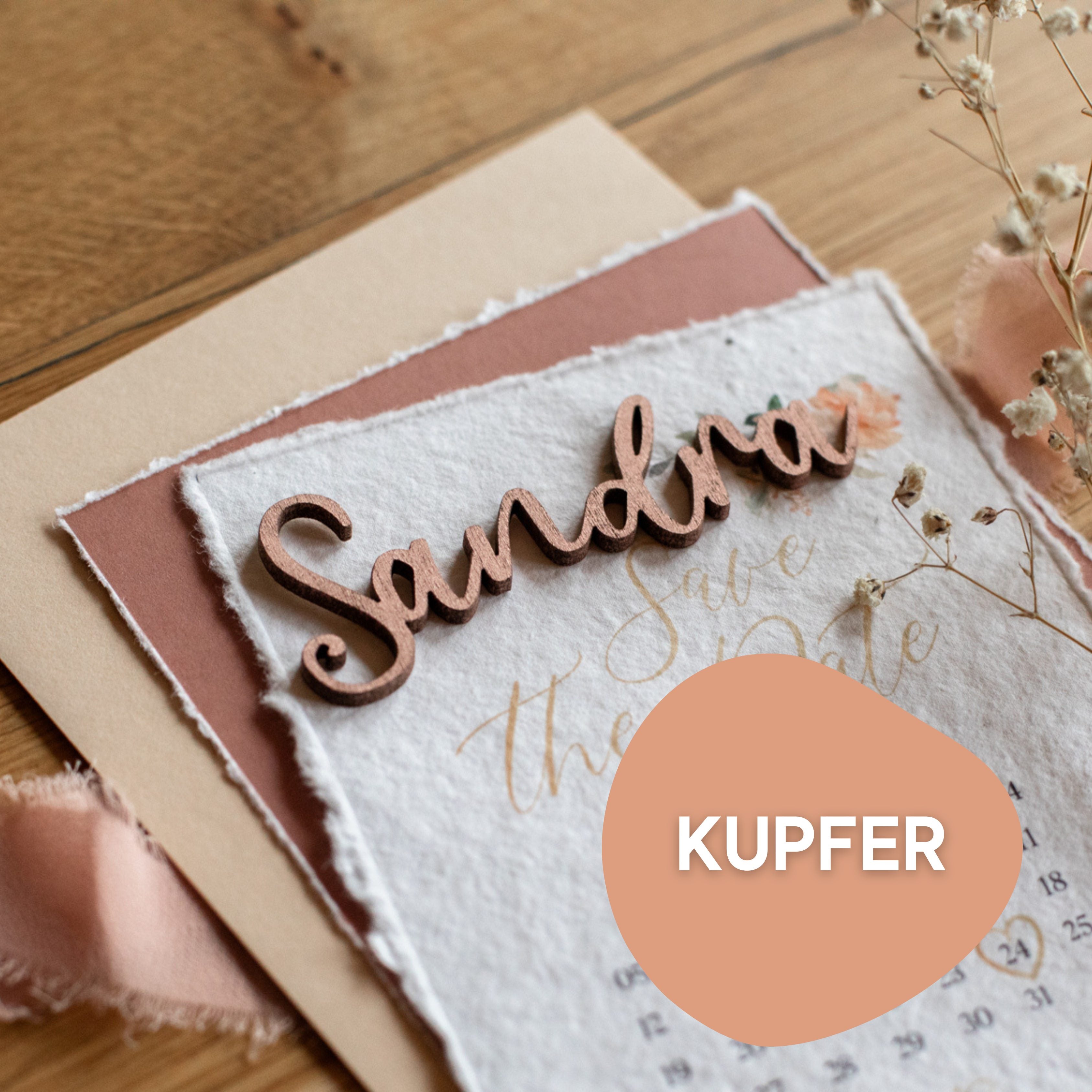 Nahaufnahme einer „Save the Date“-Karte mit einem handgefertigten, eleganten Holznamen, „Sandra“, aus Laserheldens Hochzeit Tischkarten Schriftzug aus Holz in Gold, Silber oder Kupfer, platziert auf strukturierten und pastellfarbenen Papieren.