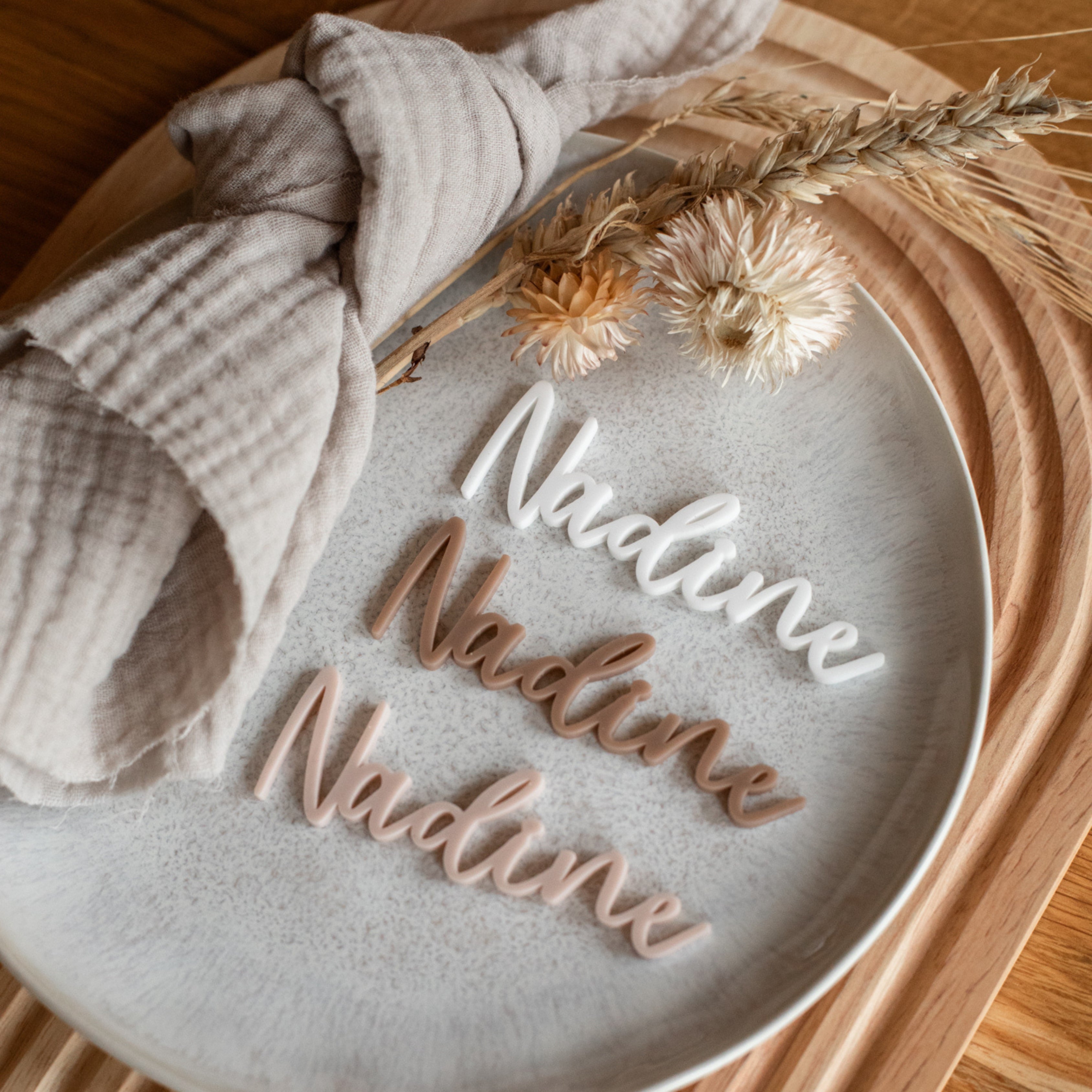 Auf einem Holztisch liegt ein grauer Teller mit drei laserhelden Namensschildern Hochzeit, personalisierten Tischkarten, einer beigen Serviette und Trockenblumen.