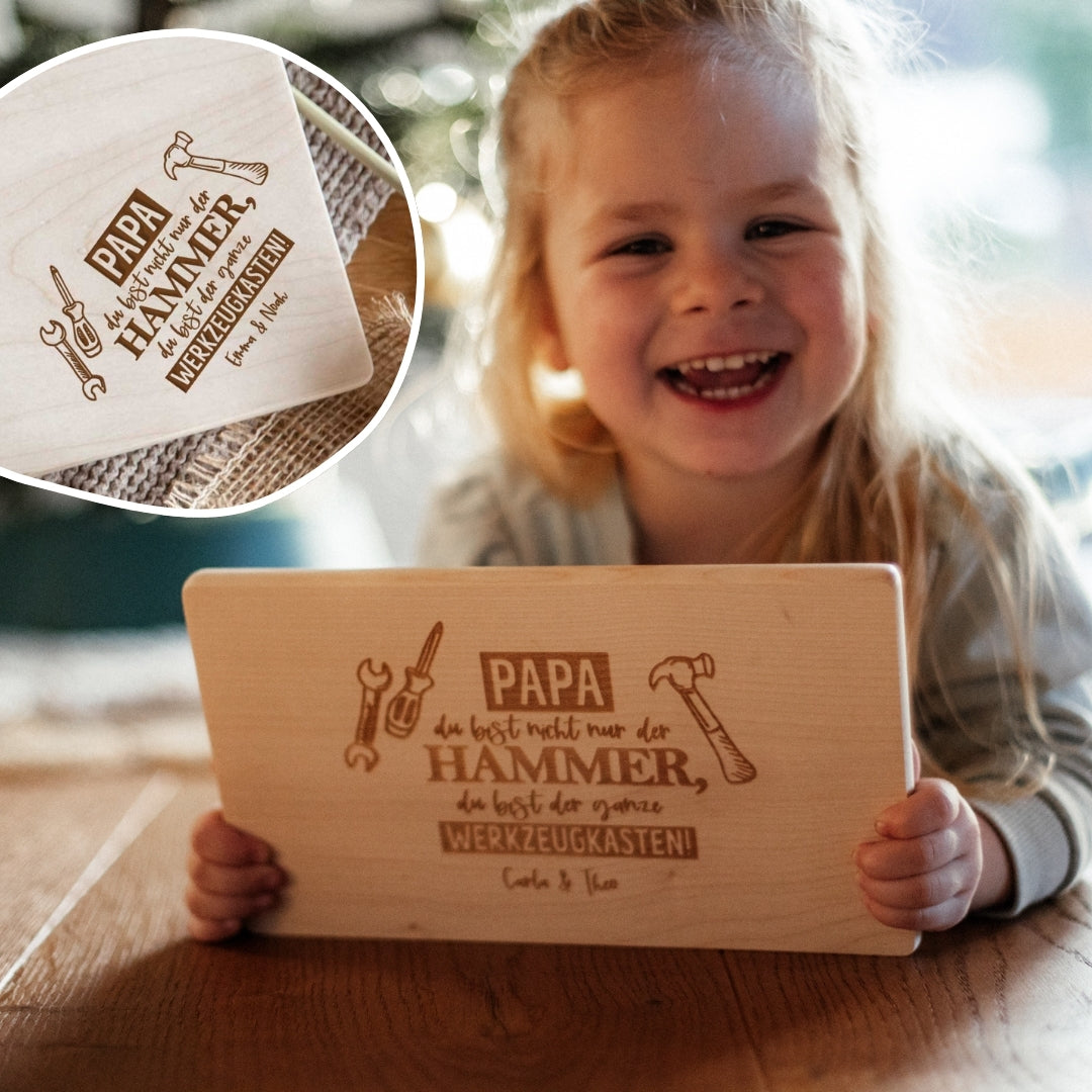 Ein lächelndes Kind hält ein laserhelden Personalisiertes Brotzeitbrett mit Gravur – Geschenk zum Vatertag für Papa, ein Holzbrett mit eingravierter Vatertagsbotschaft und Werkzeugen.