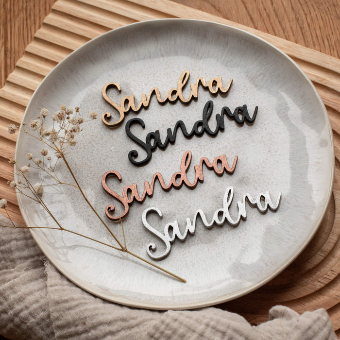 Holz-Namensausschnitte in „Sandra“, erhältlich in Gold, Silber oder Kupfer von Laserhelden, ruhen auf einem Teller mit einer getrockneten Blume und einem Tuch. Dieses charmante Arrangement dient sowohl als eleganter Holzname als auch als entzückende handgefertigte Tischdekoration.