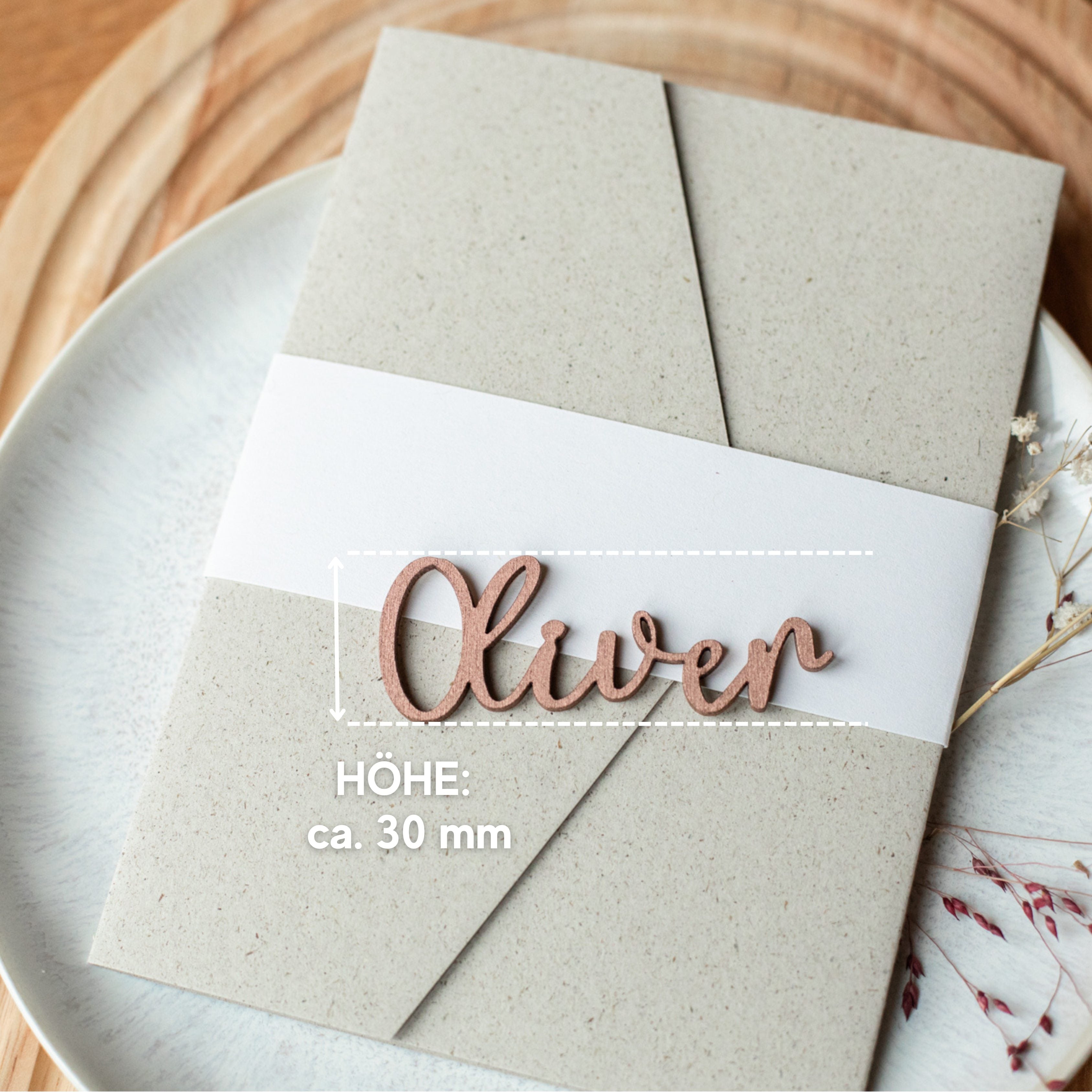 Elegant und handgefertigt: Laserheldens Tischkärtchen in Holz als Schriftzug bieten eine stilvolle Präsentation. Höhe ca. 30 mm, erhältlich in Gold, Silber oder Kupfer mit individuellem Namen wie „Oliver“ auf einem grauen Umschlag auf einem Teller.
