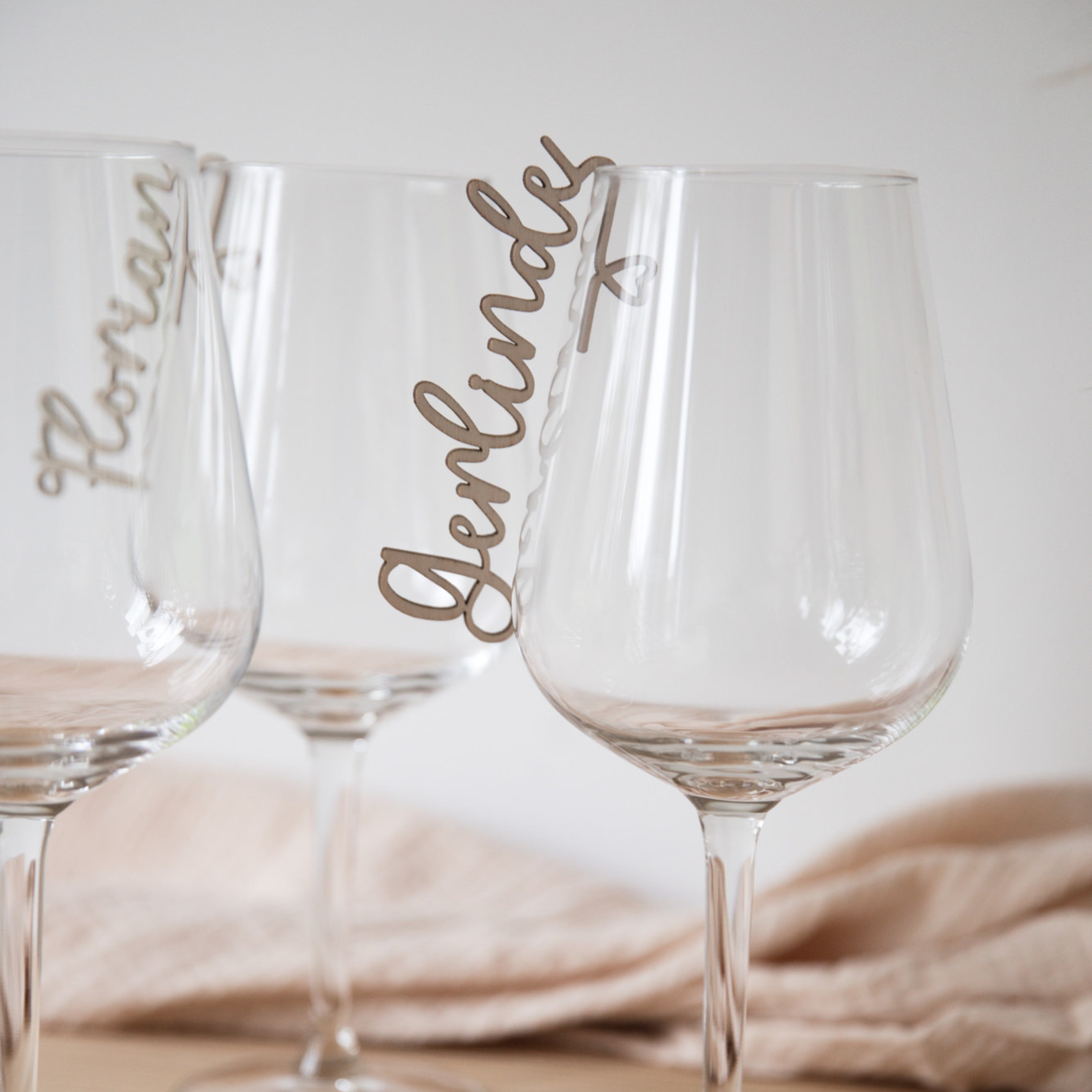 Drei klare Weingläser sind jeweils mit einem laserhelden Glasmarkierer mit Namen aus Holz verziert, der elegant am Rand hängt und sich perfekt als Hochzeitsdekoration eignet.