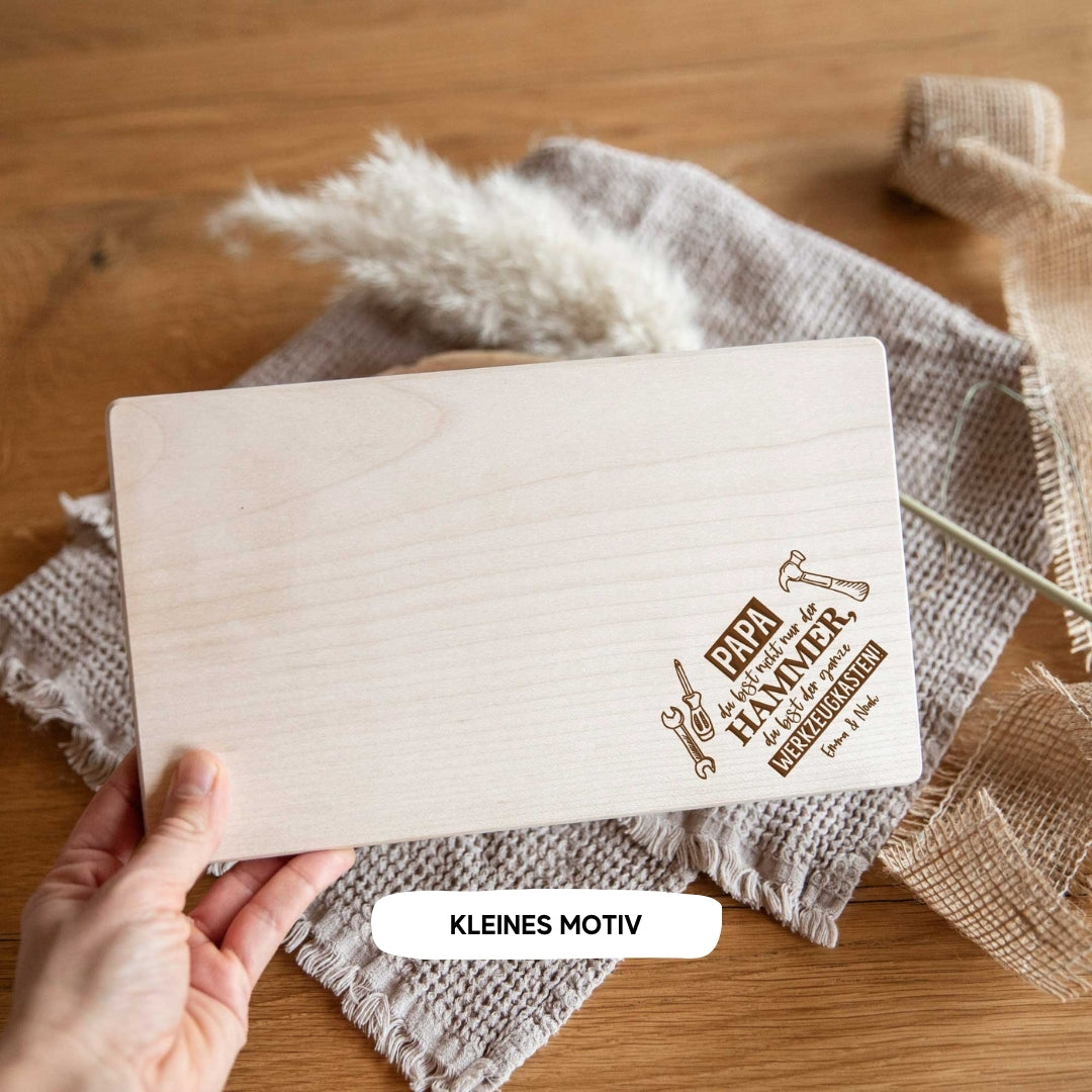 Eine Hand hält ein laserhelden personalisiertes Brotzeitbrett mit Gravur – Geschenk zum Vatertag für Papa, verziert mit Werkzeug-Gravuren und deutschem Text, auf einem strukturierten Tuch platziert.
