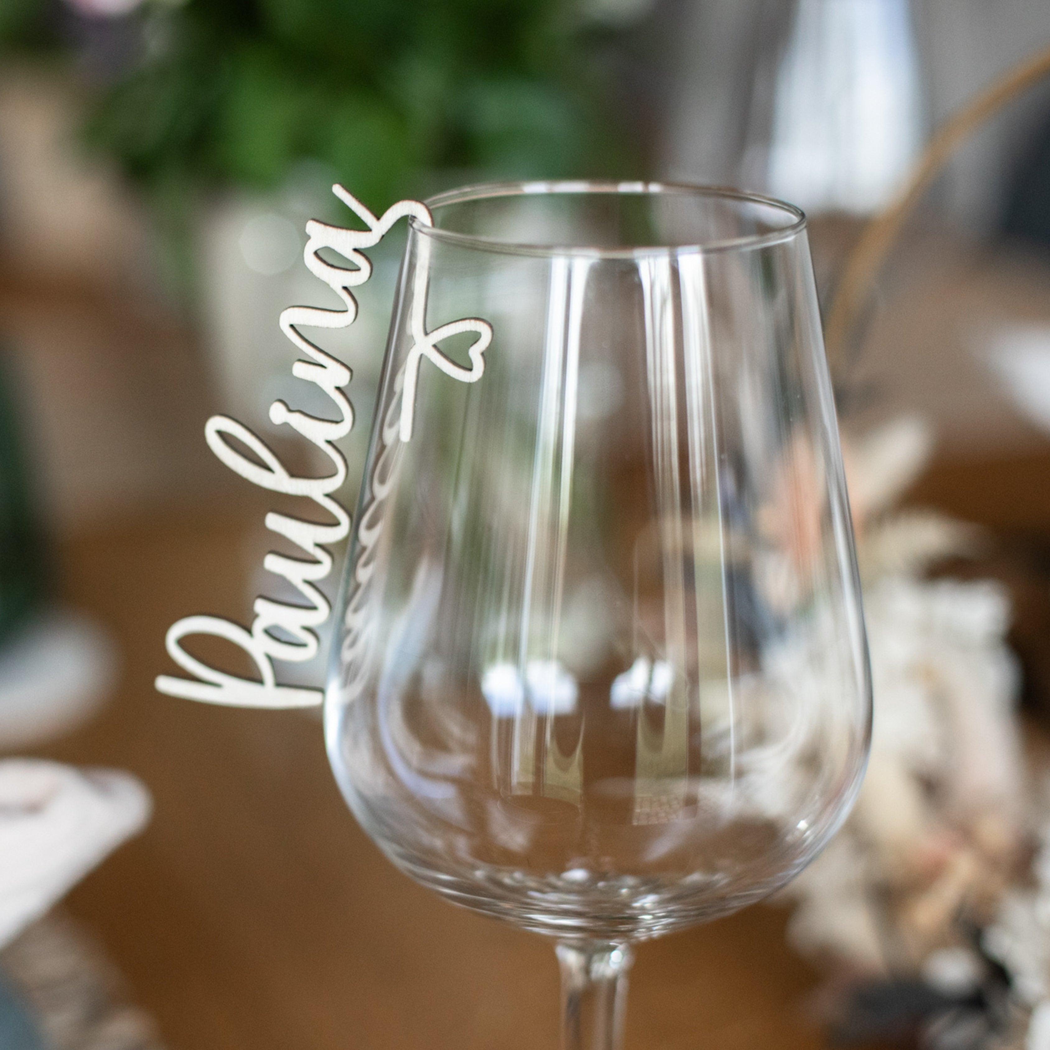 Ein Weinglas, das mit dem „Glasmarkierer mit Name aus Holz“ von laserhelden und der eleganten Aufschrift „Paulina“ verziert ist, fällt auf und unterstreicht das verschwommene Grün im Hintergrund.