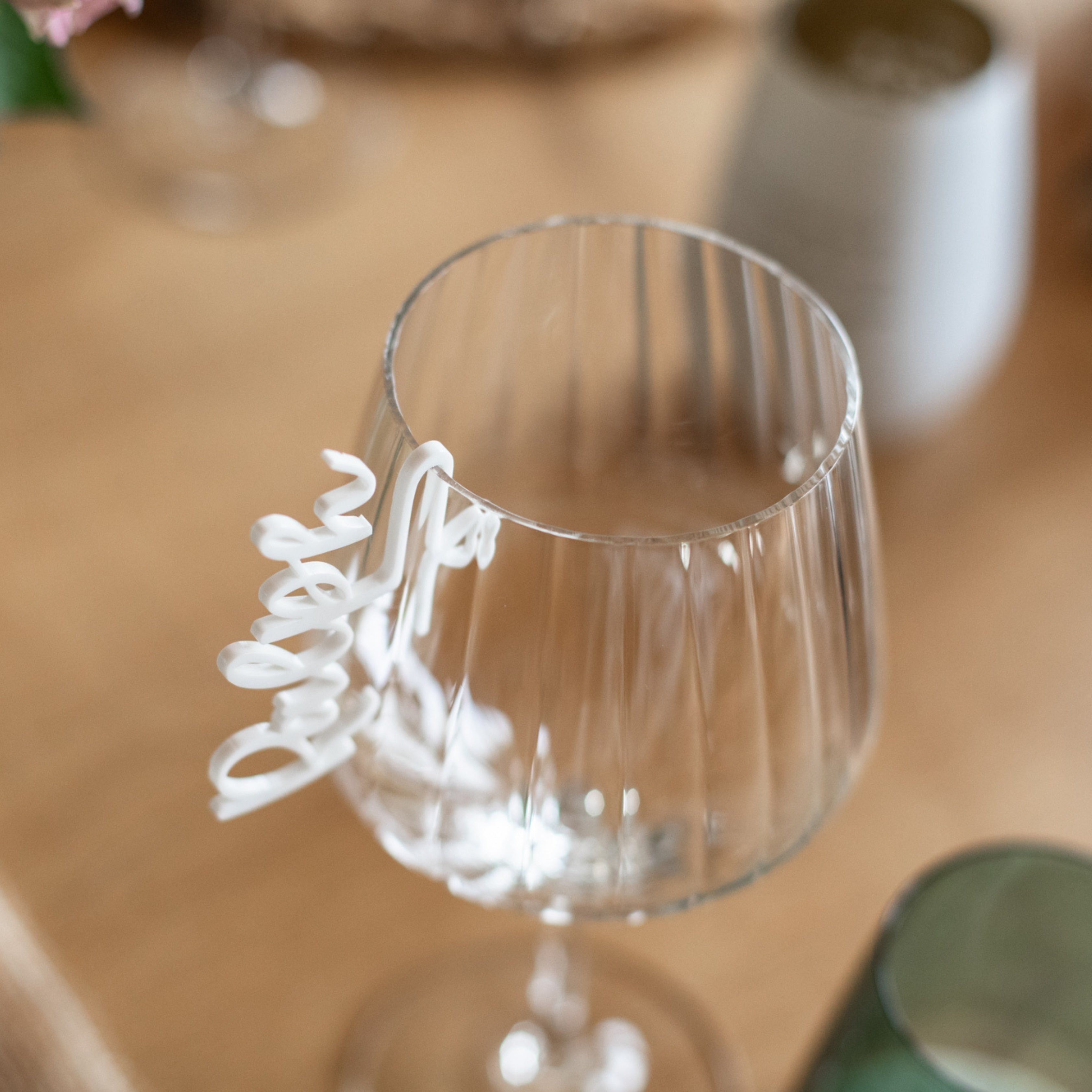 Ein Weinglas mit einem weißen, kursiven Namensanhänger von laserhelden auf einem Holztisch ist das Highlight des „Glasmarkierers personalisiert aus Acryl“, ideal für jeden Anlass.