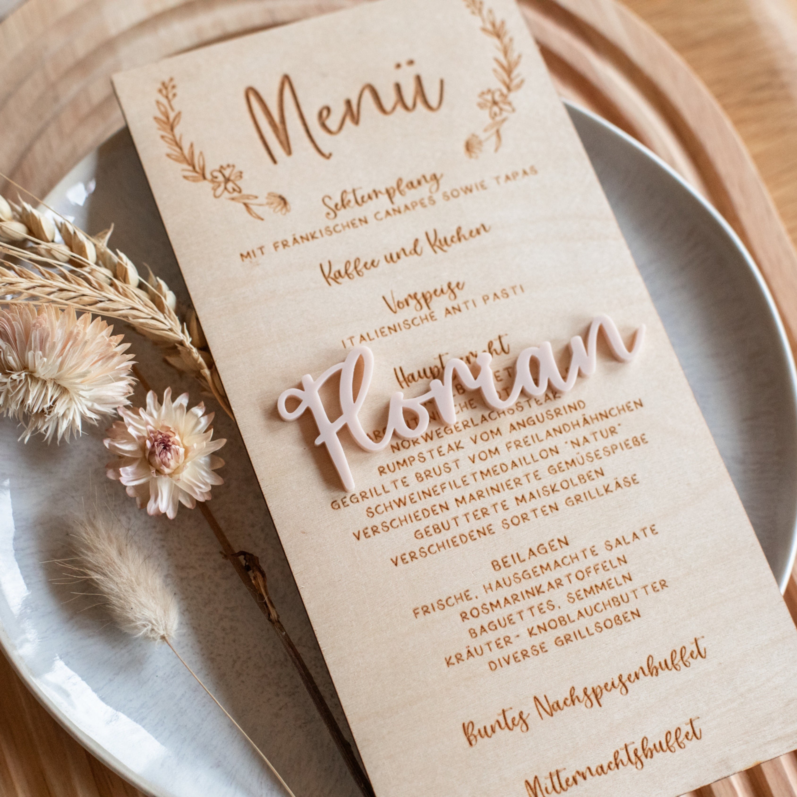 Ein elegantes Hochzeitsmenü ergänzt die Blumendekoration, wobei „Florian“ in wunderschöner Schrift auf laserhelden „Namensschilder Hochzeit“ geschrieben ist und anmutig zwischen getrockneten Blumen ruht, um jedem individuell gedeckten Tisch Raffinesse zu verleihen.