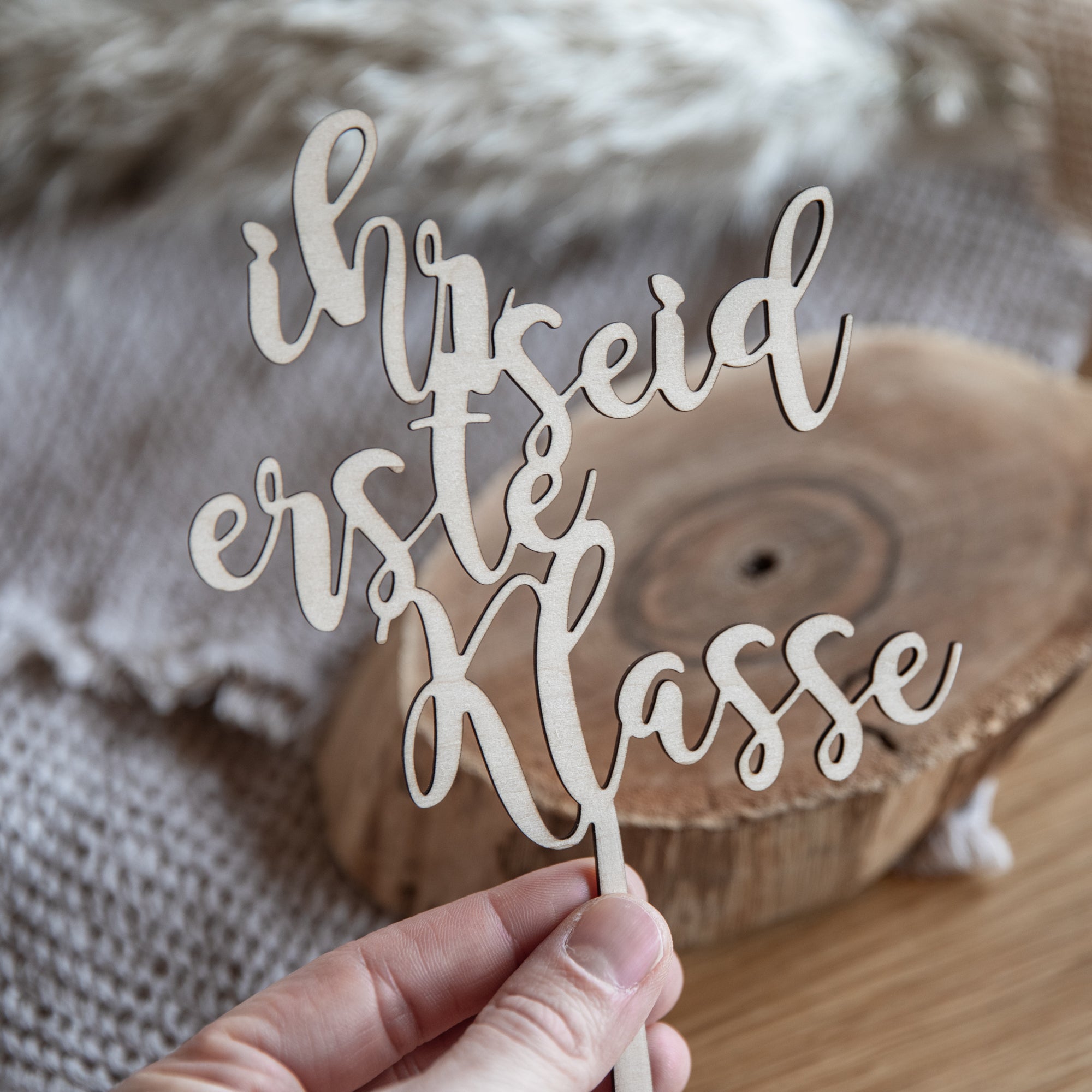 Eine Hand hält vorsichtig eine laserhelden-personalisierte Holzschrift „Ihr seid erste Klasse“ und schafft so einen einzigartigen Caketopper zur Einschulung vor einem gemütlichen, strukturierten Hintergrund.