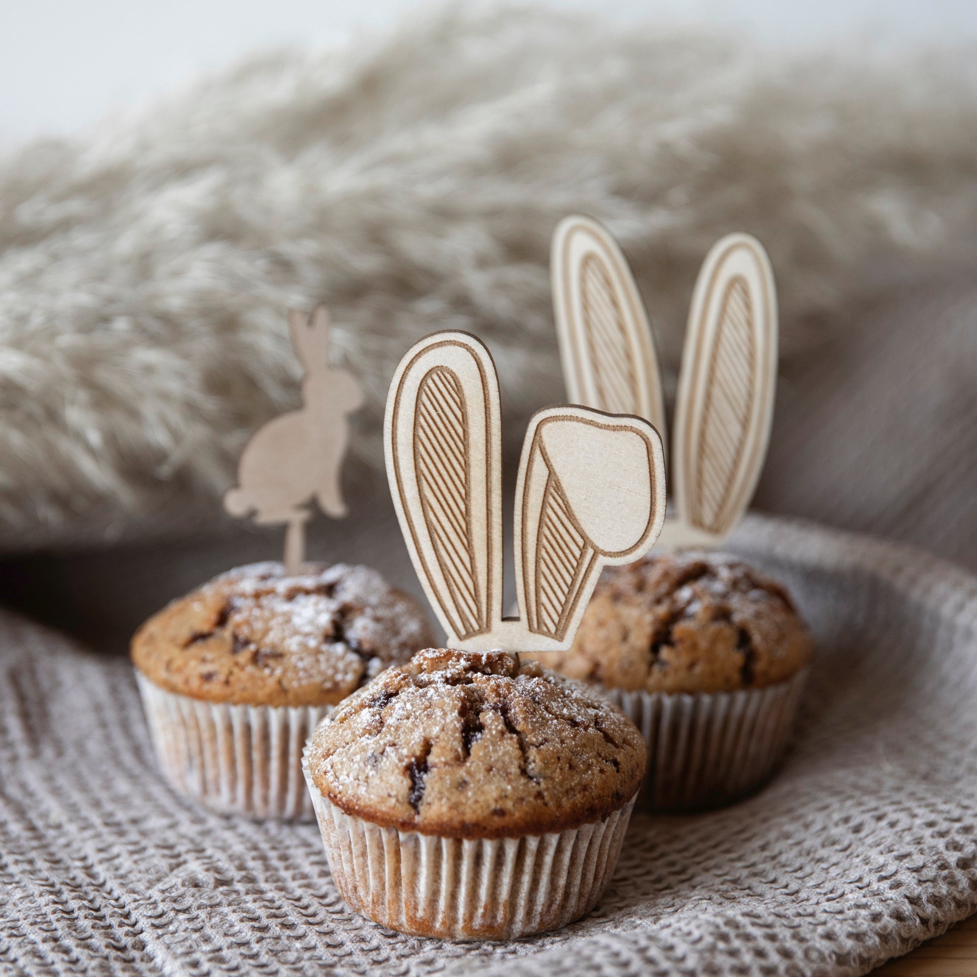 Drei Muffins sind mit laserheldens Cake-Topper/Muffinstecker - Hasen verziert, mit Hasenohren aus Papier, und mit Puderzucker bestäubt. Diese skurrilen Hasenmotive sitzen bezaubernd auf einem strukturierten Stoffhintergrund.