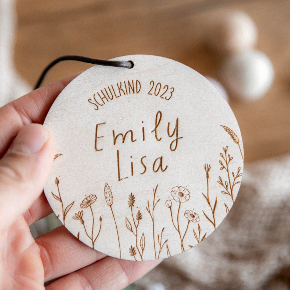 Eine Hand hält einen runden hölzernen „Schultütenanhänger zur Einschulung aus Holz“ von laserhelden mit der Gravur „Schulkind 2023 Emily Lisa“ und einem zarten Blumenmuster.