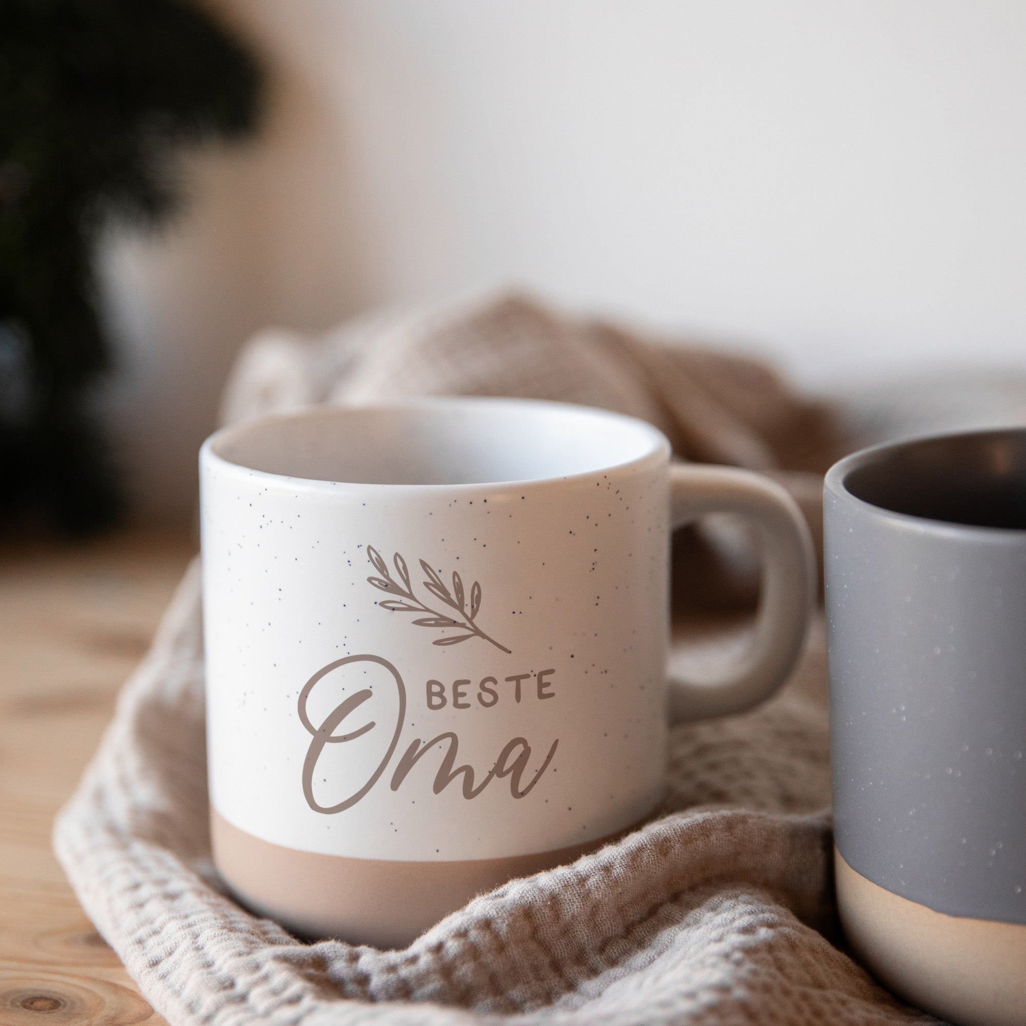 Die personalisierte Tasse aus Steingut von Laserhelden mit „Beste Oma“ und filigranem Blatt-Design ist das perfekte Geschenk, individuell graviert und stilvoll auf einer beigen Strickdecke neben einer grauen Tasse präsentiert.