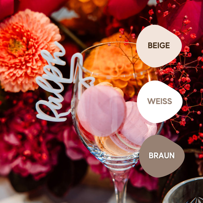 Die „personalisierten Glasmarkierer aus Acryl“ von Laserhelden schmücken elegant Weingläser und ergänzen rosa Macarons inmitten leuchtend orangefarbener und roter Blumen. Mit dem Namen „Ralph“ und Farbmustern versehen, verleihen sie als personalisierte Marker jeder Hochzeit oder Veranstaltung eine charmante Note.